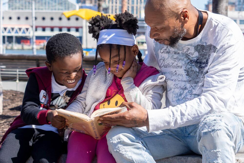 Pratt Library FY26 Annual Report  cover photo.- a family reading in Baltimore.