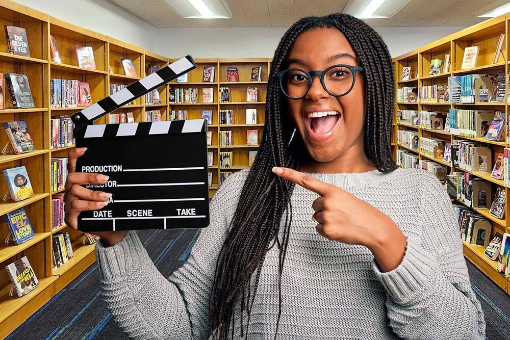 book trailer contest - excited person pointing to a filming clapboard with a background of Pratt Library books for teens