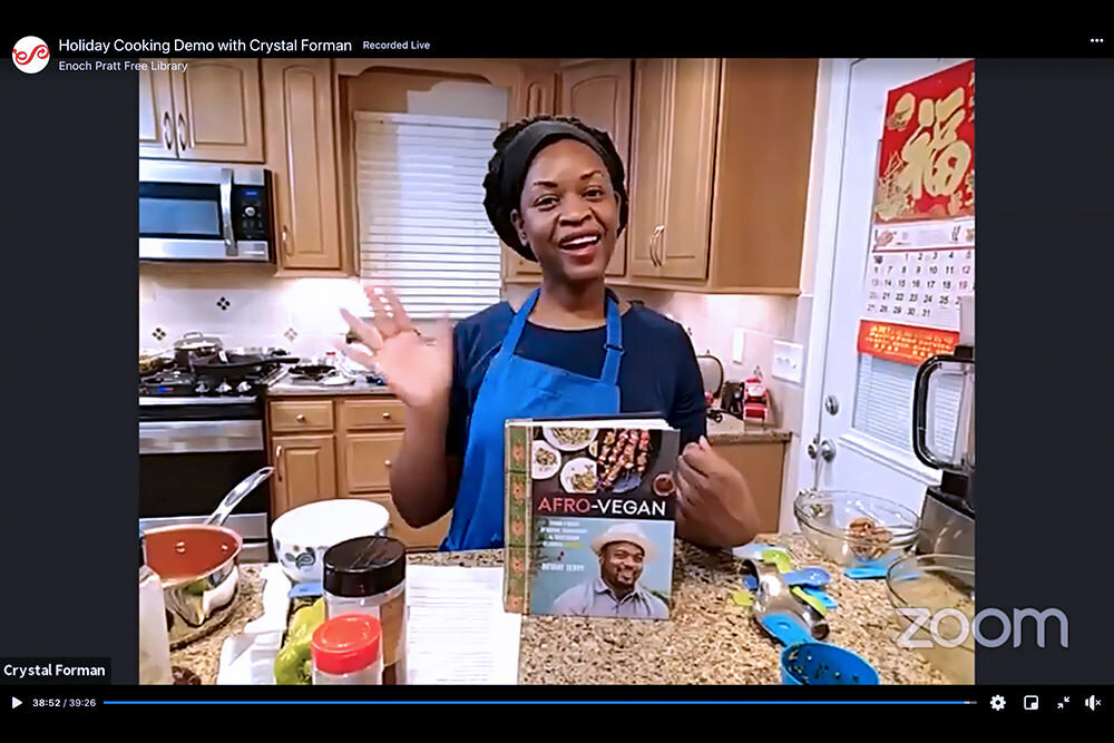 Cooking demo on Zoom - Holiday Cooking with Crytsal Foreman waving in a kitchen and the Afro Vegan book