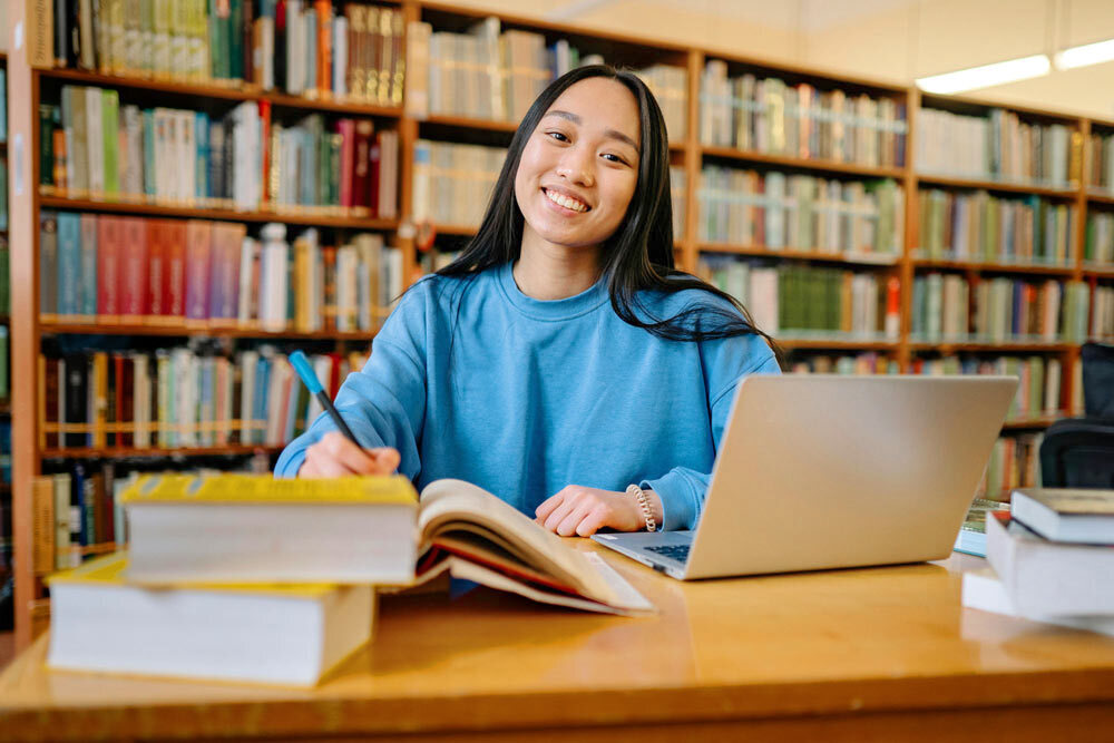smiling woman at a wooden library table with books, pen, laptop