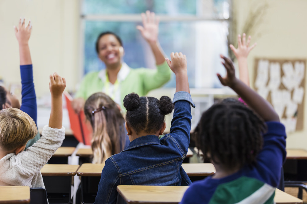 teacher and kids with hands raised in a classroom