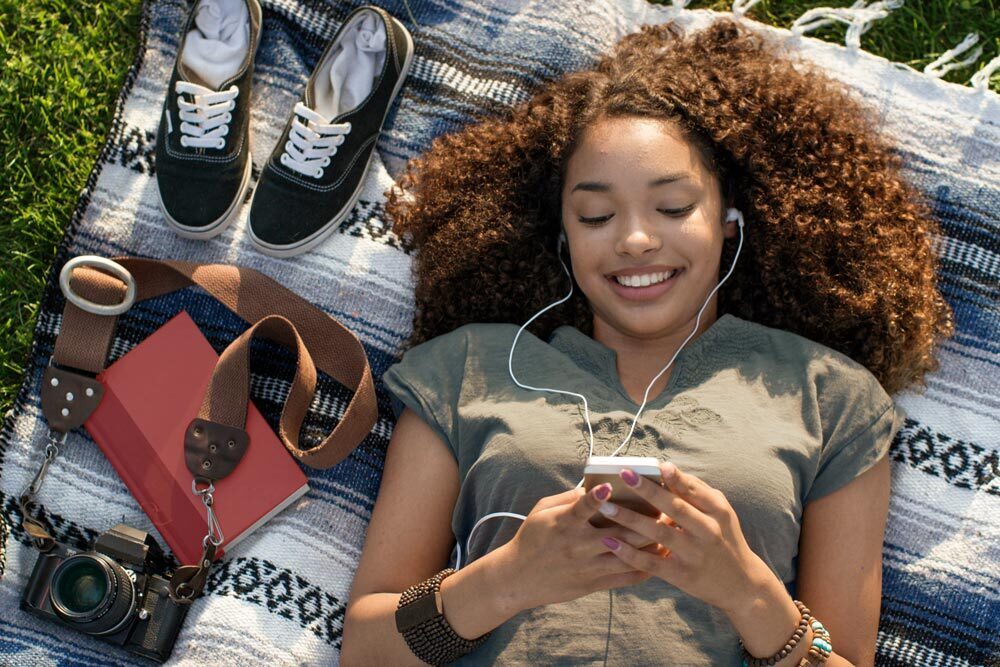 a teen girl lying on a outdoor blanket with a book, camera, audio device, headphones, and shoes