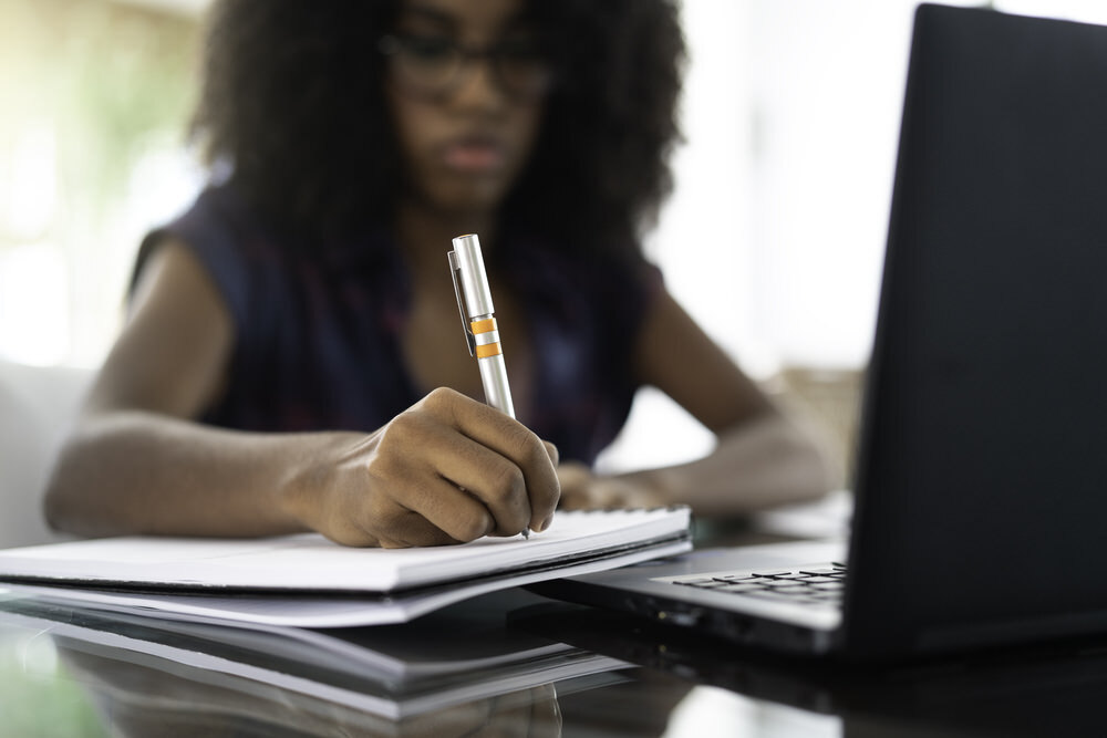 person writing with pen to notebook and a laptop