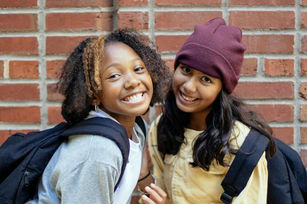teens girls smiling with backpacks by a brick wall