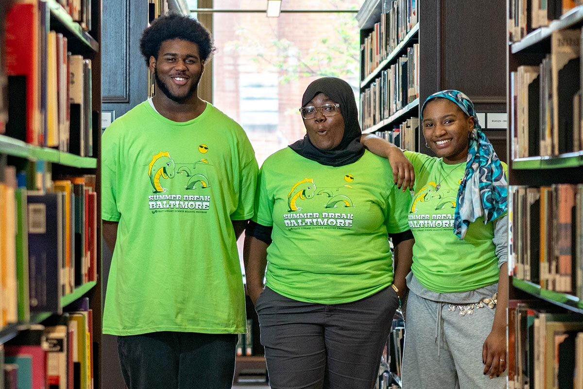 3 teens wearing Summer Break Baltimore 2026 t-shirts by bookshelves.