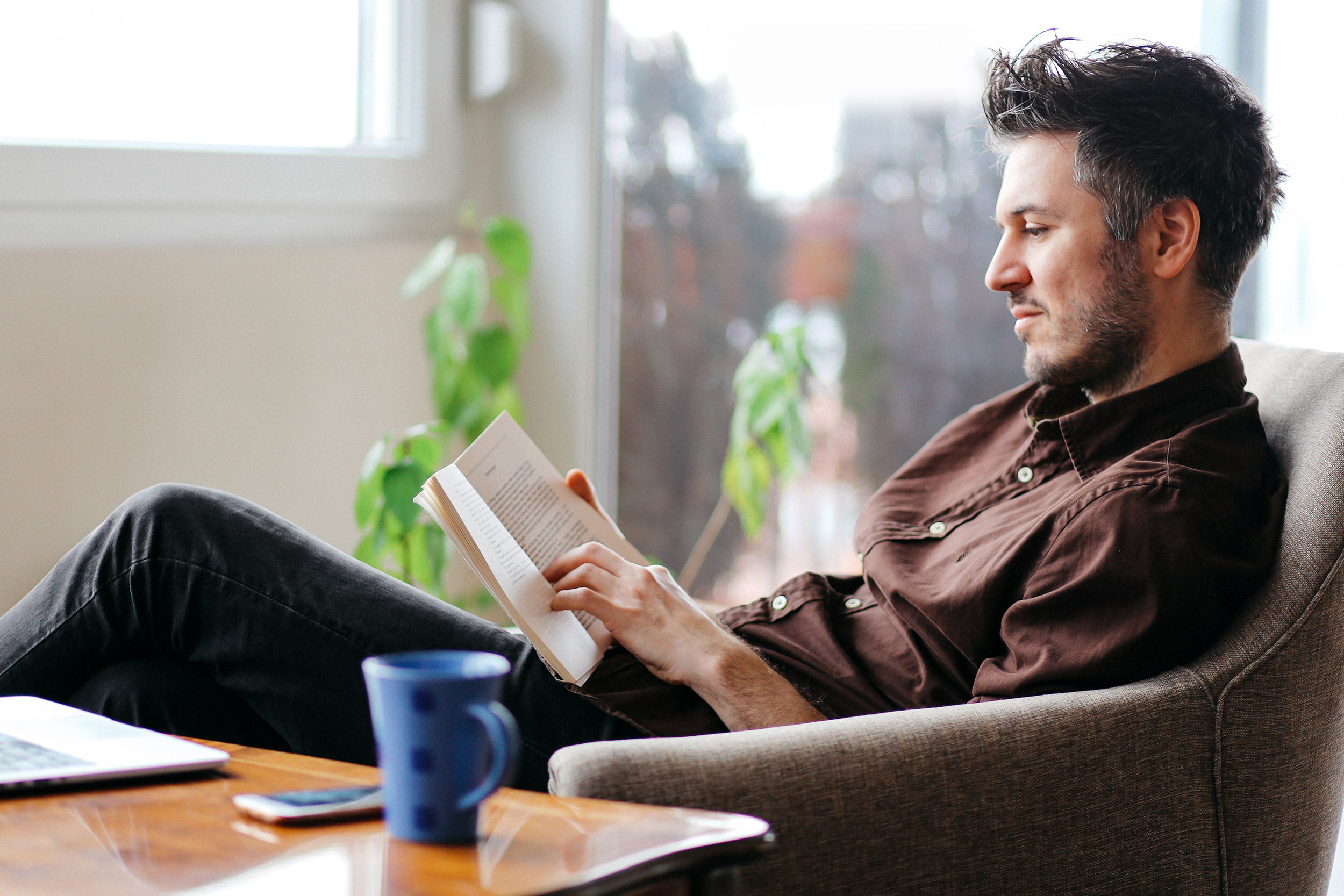 adults - a man reading a book at home with coffee and laptop