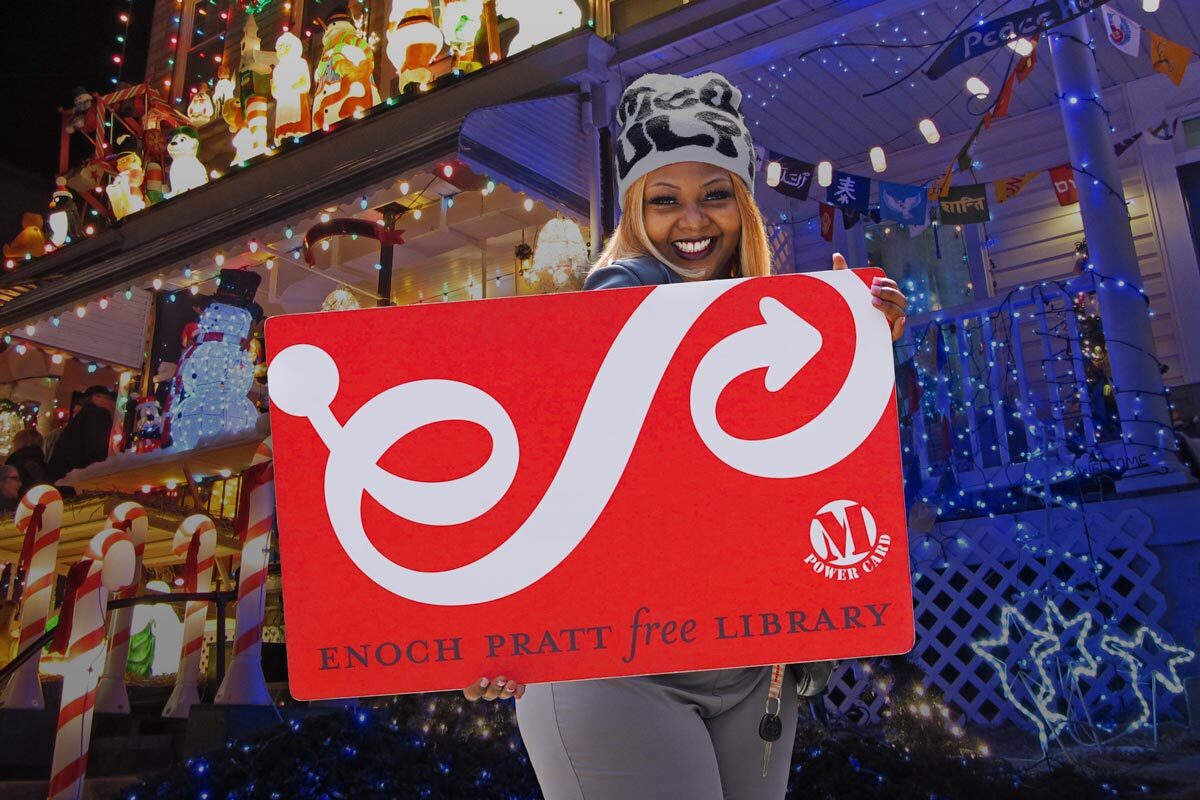oversized Pratt Library card held by a smiling woman with Baltimore holiday lights in the background