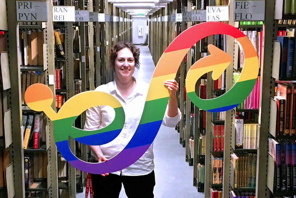 Pratt staff with a rainbow LGBTQ Pride Pratt logo sign in the book stacks