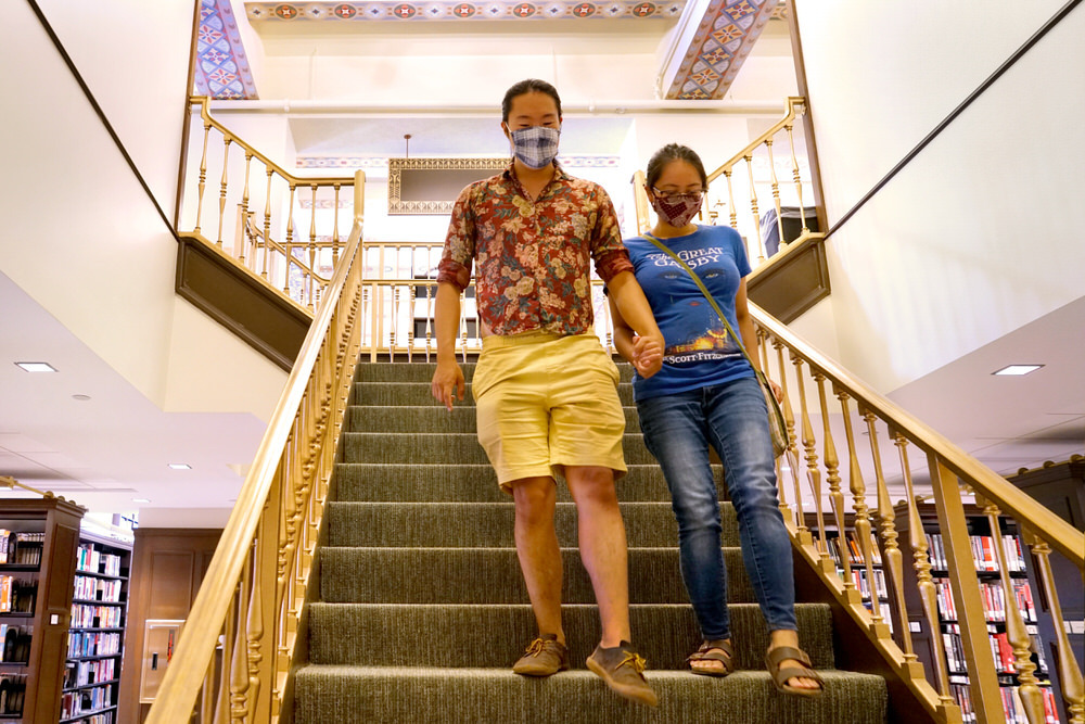 Couple in Central Library, walking down stairs, smiling, wearing face masks. One is wearing a Great Gadsby book cover shirt.