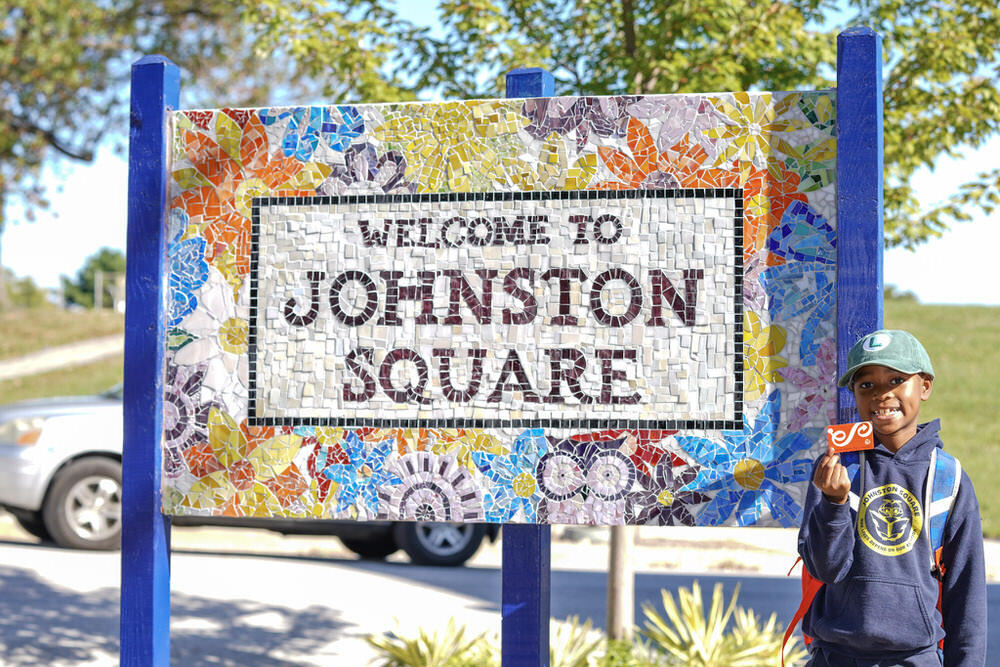 child with a Pratt library card by the Johnston Square Branch sign