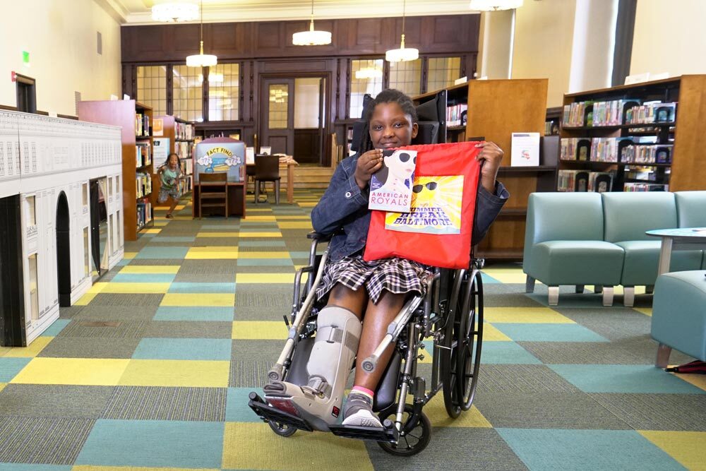 girl using a wheelchair in the Central Library's Children's Department, holding up a free Summer Break Baltimore bag and book