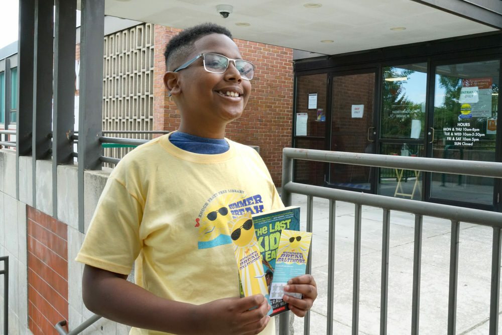 boy wearing a Summer Break Baltimore shirt, holding a free book, bookmark, and scratch-off card