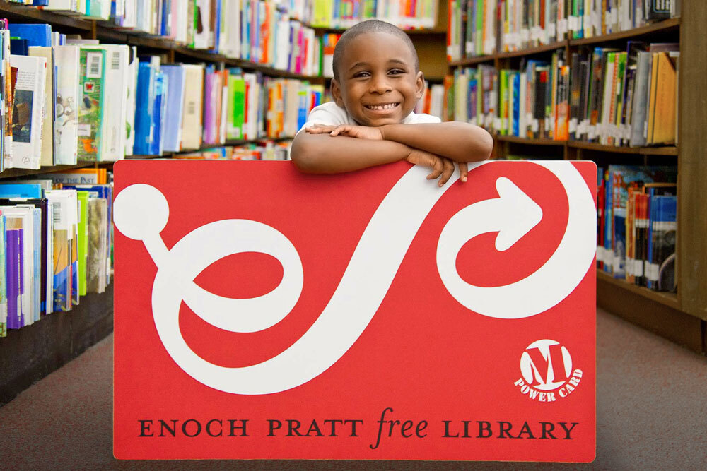 smiling boy with an oversized Pratt Library card in the bookshelves