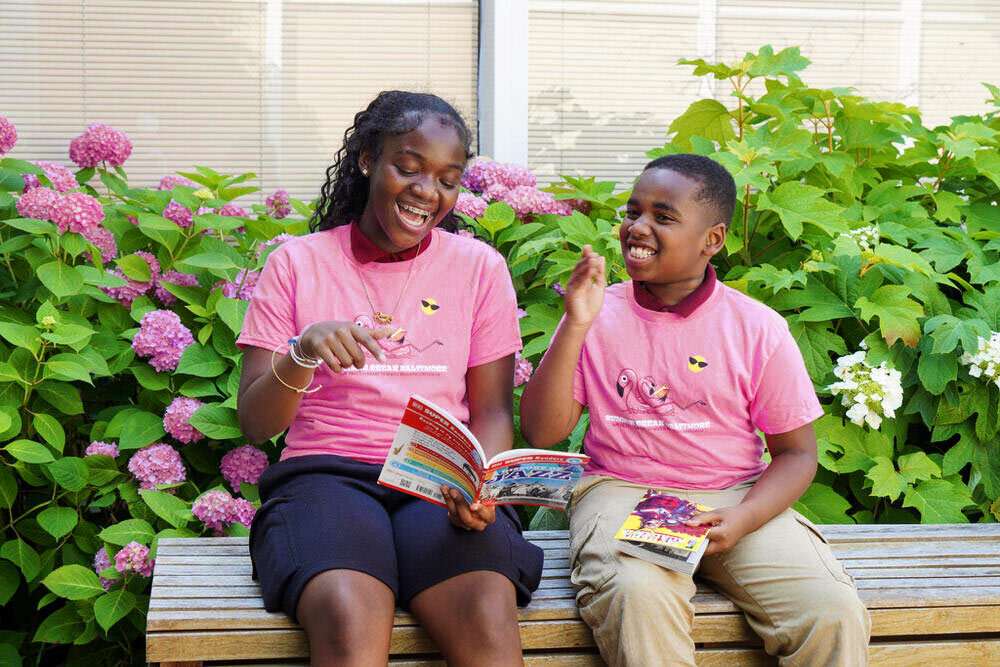 two kids with books in Summer Break Baltimore t-shirts outside at the Hamilton Branch