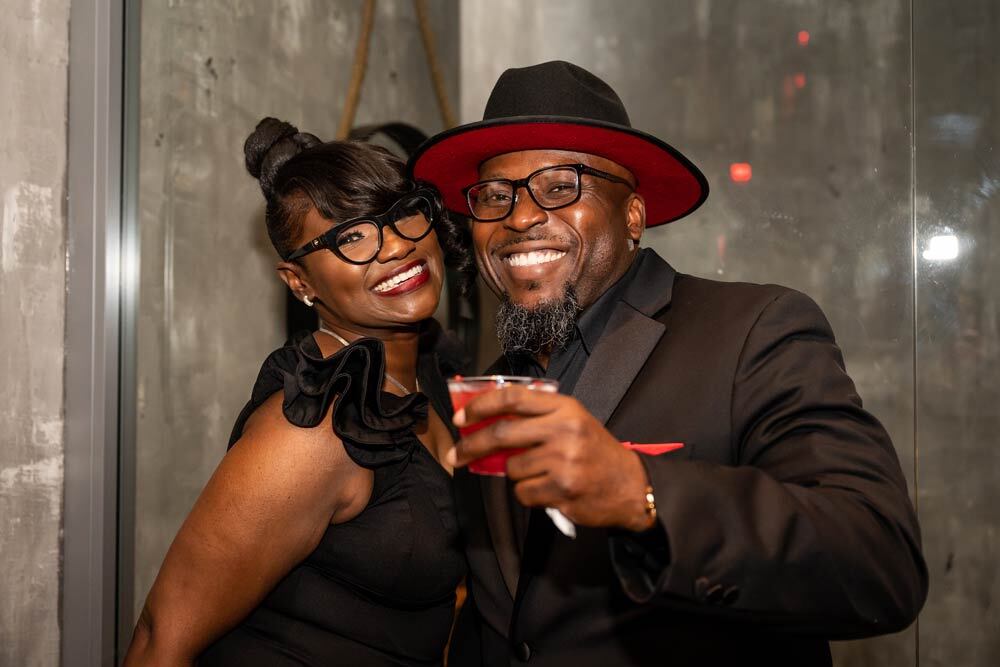 a couple smiling at the 2022 Black & White Party, with black clothes, a red-accented black hat and red drink
