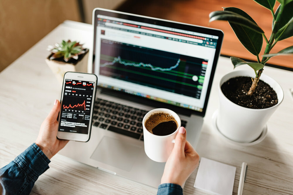 stock transfers - person at a desk with investment chart screens, plants and coffee