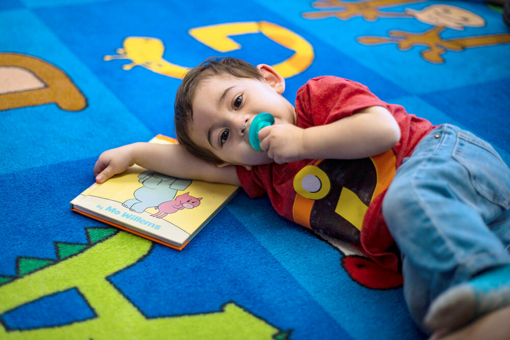 young boy with pacifier with book on rug at Patterson Park storytime