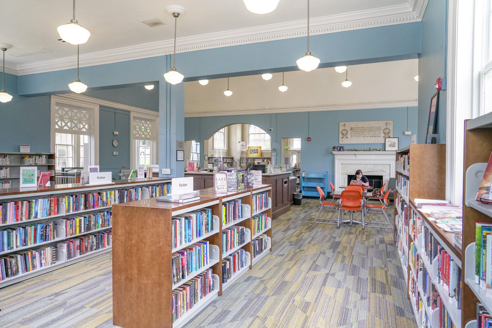 Hampden interior with low bookshelves, windows, lights, and an  adult reading at the back