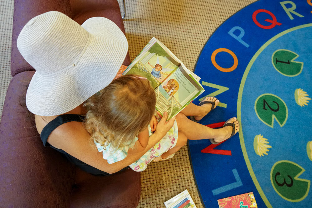 Roland Park - woman wearing a hat reading a book to young child by a colorful rug